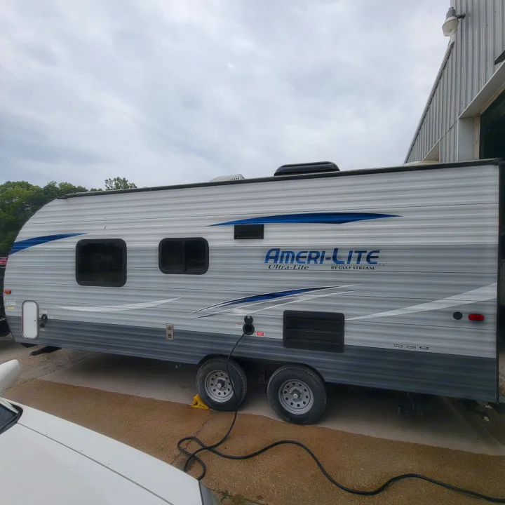 Light gray Ameri-Lite travel trailer, perfect for an rv rental, parked on asphalt. Features blue stripes & is plugged into power.. Gulf Stream Ameri-Lite 2018