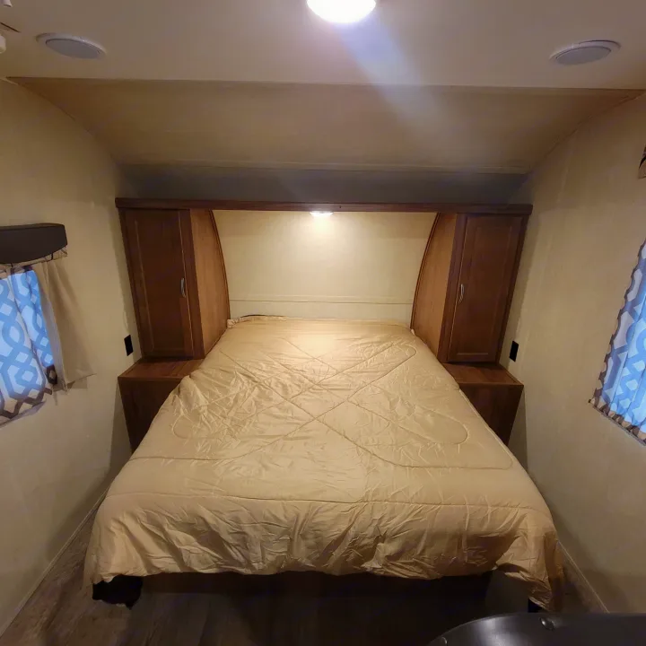 RV rental bedroom: a spacious bed with light comforter, flanked by cabinets, under bright ceiling lights. Cozy travel space.. Gulf Stream Ameri-Lite 2018