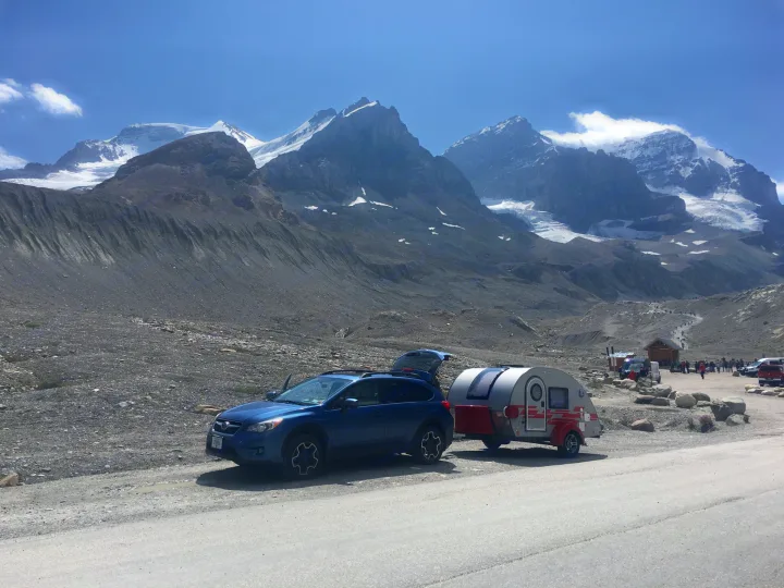 Scenic rv rental adventure: a blue SUV tows a teardrop trailer amidst vast rocky terrain and majestic snow-capped mountains.. nuCamp T@G 2018
