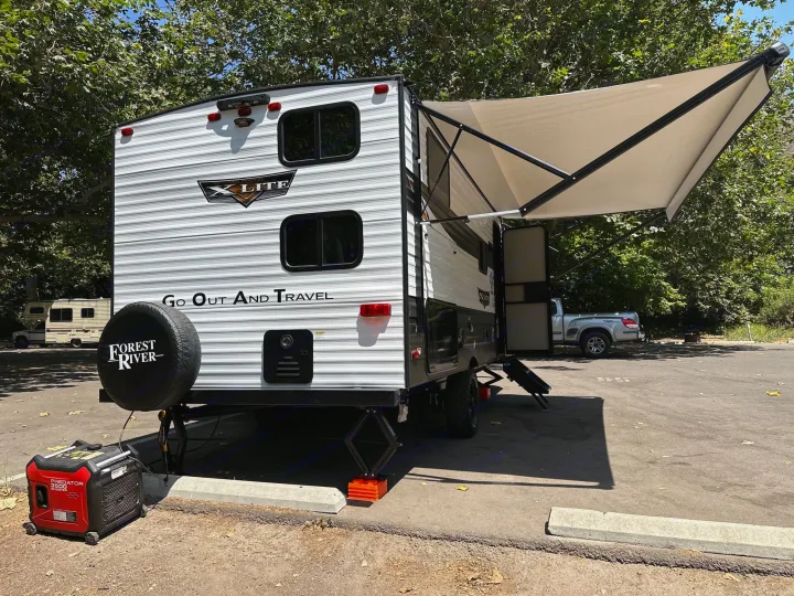 A white travel trailer RV rental with an extended awning, a red generator, and leveling jacks, perfect for camping.. Forest River Wildwood Platinum 2024