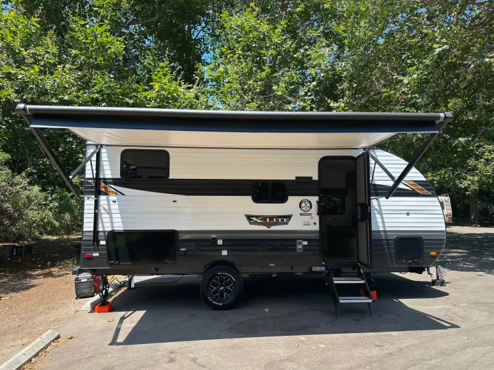 Explore the outdoors! This modern rv rental travel trailer, with its awning extended, is parked at a shaded campsite.. Forest River Wildwood Platinum 2024