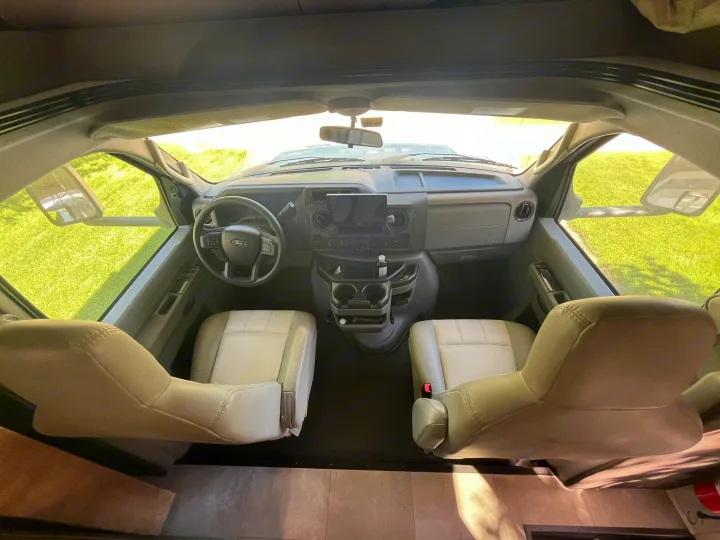 Inside an RV rental, the front cabin features light-colored driver/passenger seats and dashboard, looking out to green grass.. Coachmen Leprechaun 2021