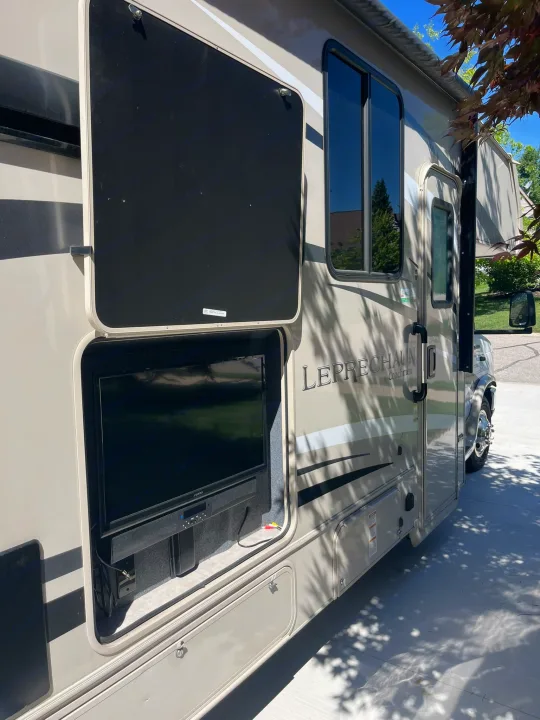 A beige **RV rental** with an open side compartment displaying an outdoor TV and soundbar, ideal for camping fun.. Coachmen Leprechaun 2021