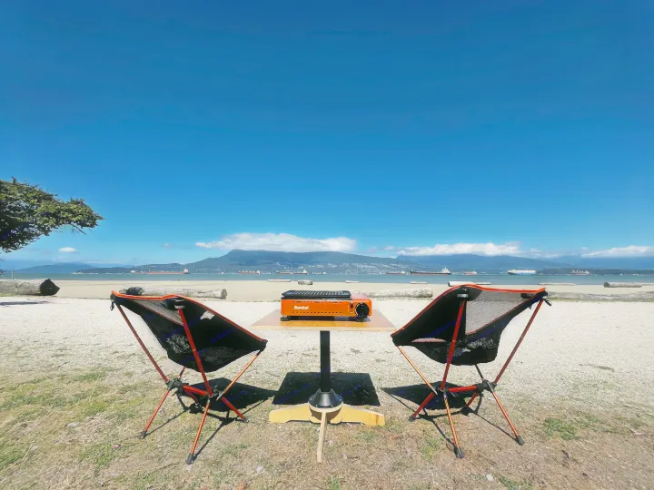 Beachside RV rental stop: Portable camping chairs, table, and grill set up by the ocean with ships and mountains.. Dodge Grand Caravan 2014