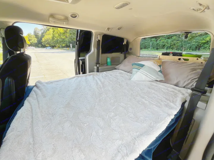Inside a cozy minivan, converted for an RV rental. Features a comfy bed, pillows, and an outdoor view of a sunny parking lot.. Dodge Grand Caravan 2014