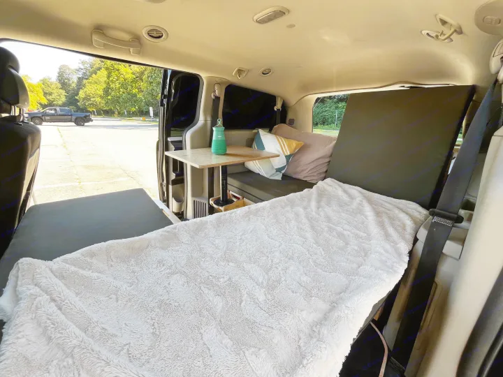 RV rental: Inside a cozy van conversion, featuring a bed with a fluffy blanket, table, and outdoor view.. Dodge Grand Caravan 2014