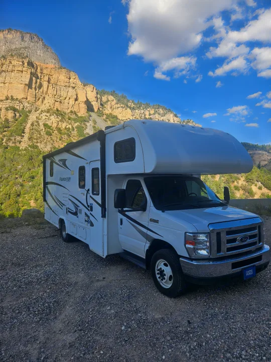 White RV rental parked on a gravel road, with tall rocky mountains and a blue sky with clouds in the background.. Forester Forester Motorhome 2020