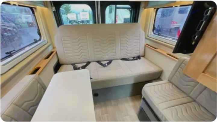 This RV rental interior features comfy bench seating with seatbelts, a functional table, and large windows revealing a rainy day.. Coachman Nova 2024
