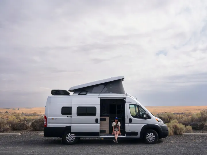 Woman relaxing in an rv rental camper van with pop-top roof, parked in a wide, dry landscape.. Other Other 2022