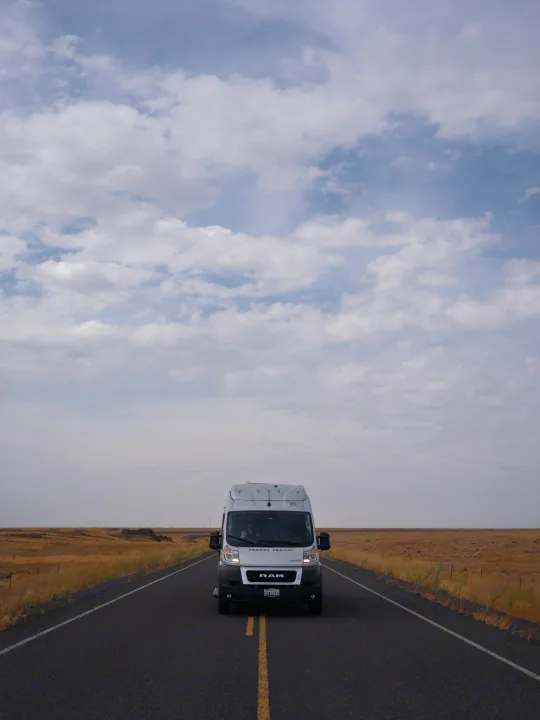 A white RV rental van drives down a straight, empty road through vast, dry fields under a cloudy sky.. Other Other 2022