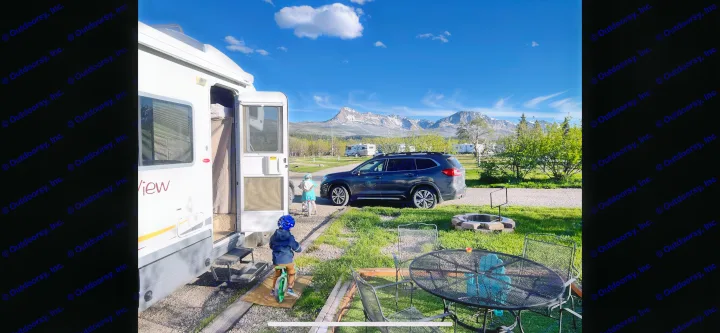 An RV rental is parked at a scenic mountain campground. Two kids on bikes explore, with a dark SUV and fire pit nearby.. Winnebago View 2007