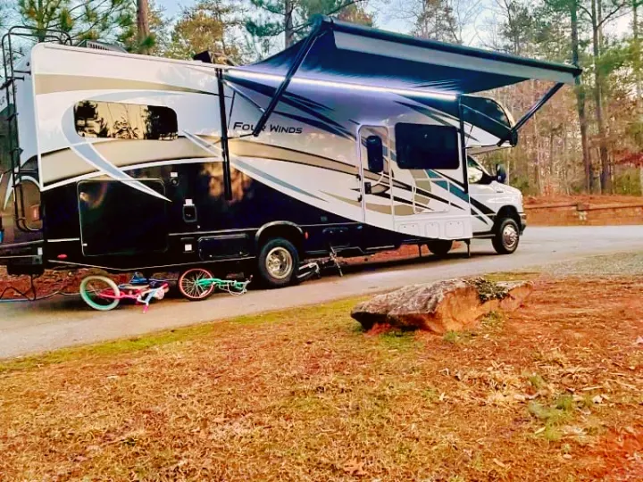 Spacious RV rental with illuminated awning parked on a paved site. Children's bikes are on the ground, perfect for family camping trips.. Thor Four Winds 2022