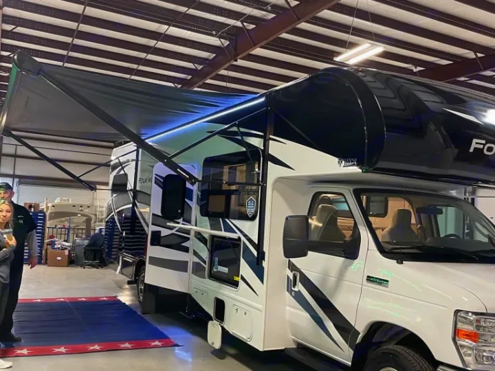 A white Class C RV with black graphics and an extended dark awning with bright LED lights is parked indoors, ready for RV rental.. Thor Four Winds 2022