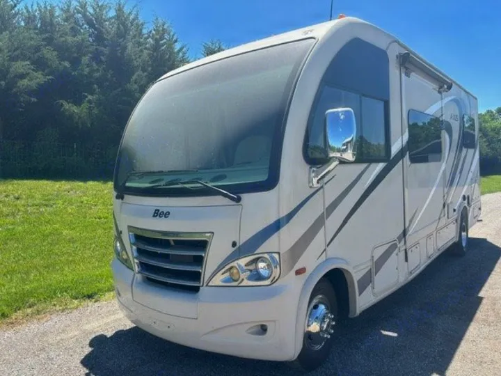 Light-colored RV motorhome available for rv rental, with gray graphics, parked on gravel under a clear sky.. Thor Axis 2016