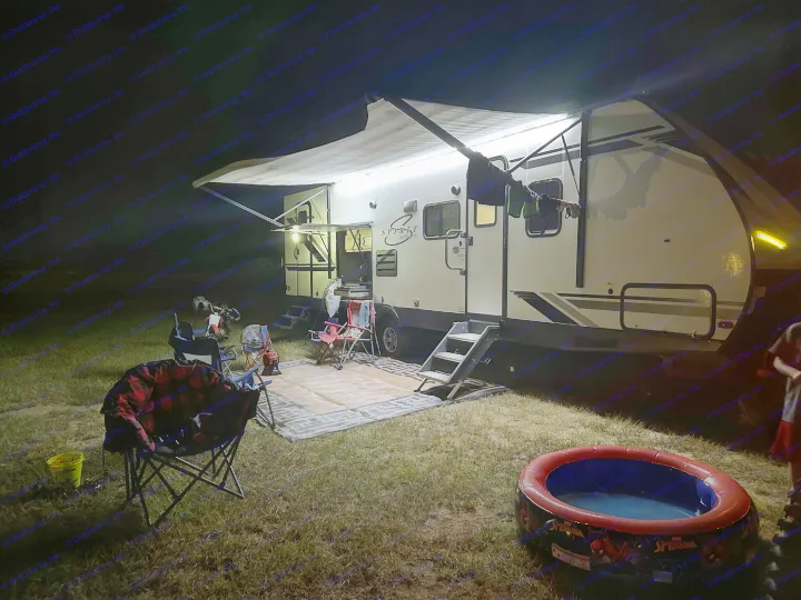 Night camping with an RV rental, illuminated by awning lights. Outdoor rug, chairs, and a small pool complete the cozy setup.. Coachmen 2021 Coachman Sprinter Campfire Edition 2021