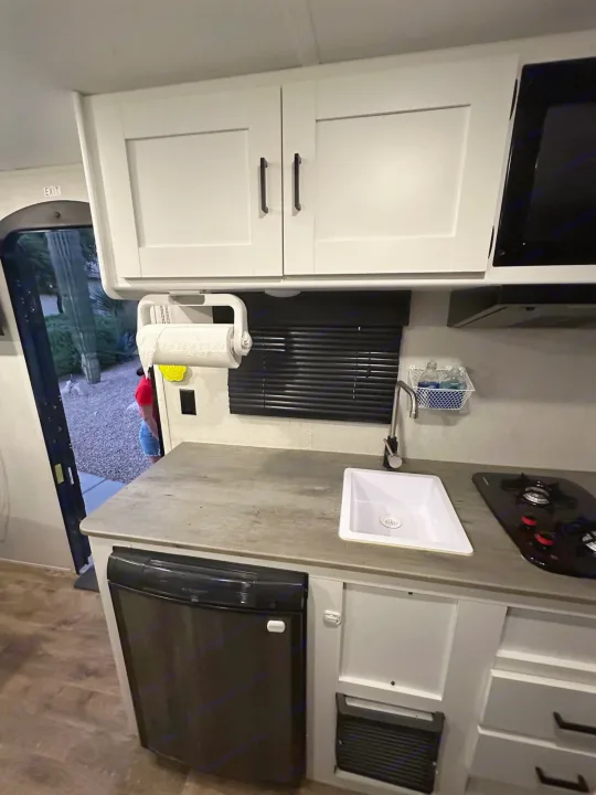 Inside an RV rental: a compact kitchen with white cabinets, sink, stove, and small fridge. An open exit door shows outside.. Keystone RV Hideout 2021