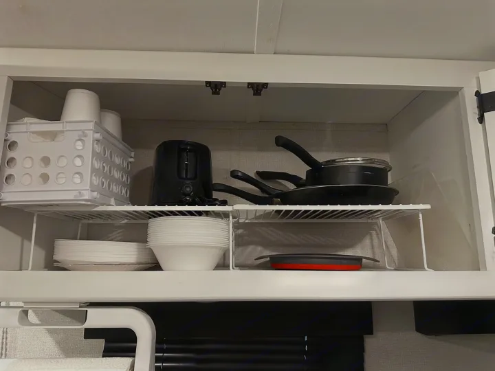 RV rental storage: White cabinet with wire shelves holding kitchen pots, pans, cups, plates, and a black toaster.. Keystone RV Hideout 2021