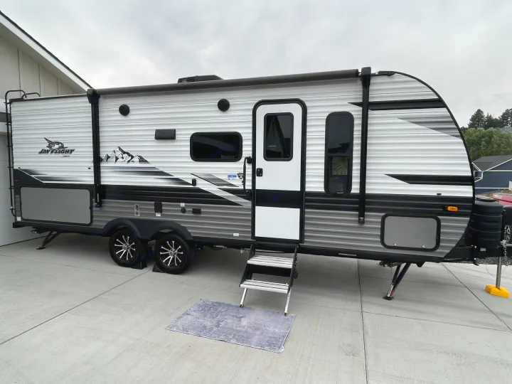 Side view of a gray & white Jay Flight travel trailer on a concrete driveway, featuring steps & awning. Ready for an RV rental.. Jayco Jay Flight 2024