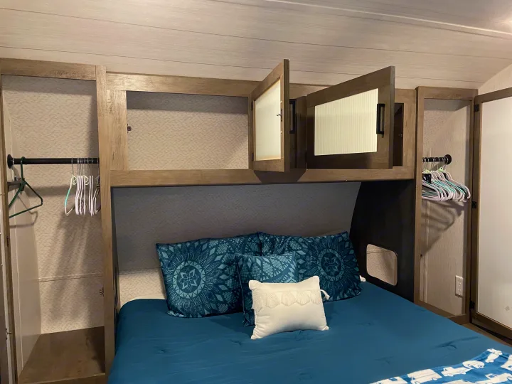 Compact RV rental bedroom features a blue bed, overhead storage, and side wardrobes with hangers, maximizing space.. Forest River Wildwood 2022