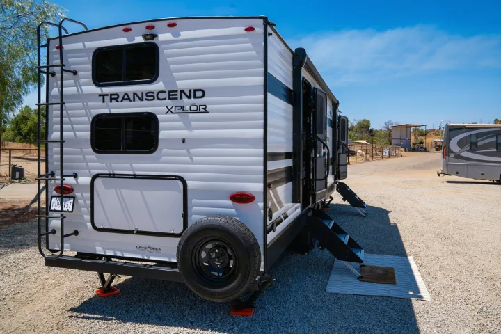 This white Transcend Xplor travel trailer RV rental is on gravel. It features a rear ladder, spare tire, and extended side steps.. Grand Design Transcend Xplor 2024