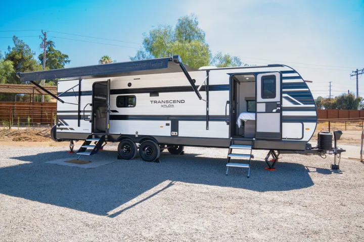 This RV rental travel trailer is fully set up on a gravel lot, with its awning extended and doors open.. Grand Design Transcend Xplor 2024