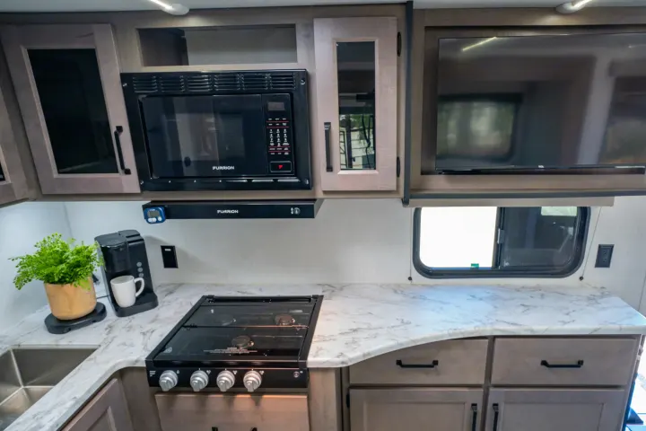 Spacious RV rental kitchen features microwave, stove, sink, and TV with marble countertops & coffee maker setup.. Grand Design Transcend Xplor 2024