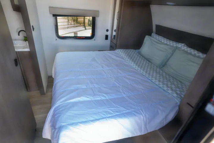 This RV rental bedroom features a light blue bed, green checkered sheets, a compact sink, and a window with an exit sign.. Grand Design Transcend Xplor 2024