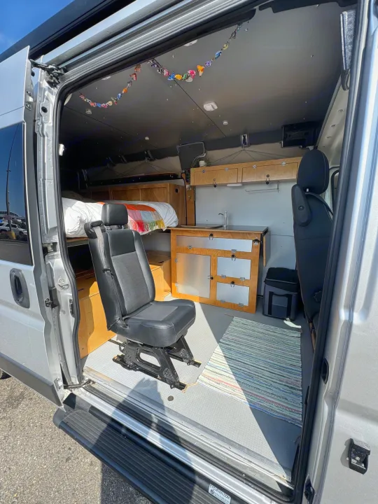 Spacious RV rental camper van interior. Features a swivel seat, bed, & kitchenette, viewed through the open side door.. Ram Promaster 2500 2021