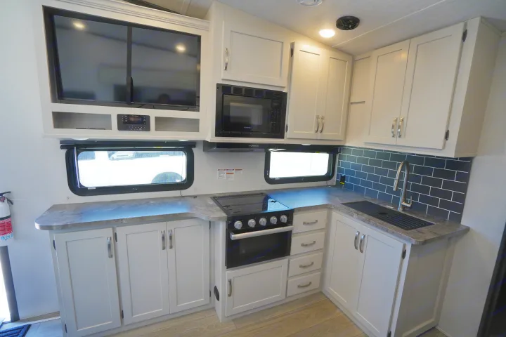 Spacious RV rental kitchen features light cabinets, dark countertops, a stove, microwave, sink, and TV.. Keystone Cougar 2024