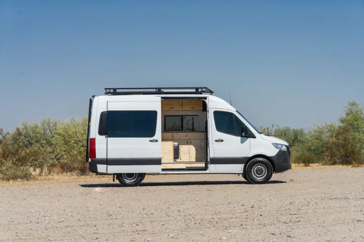 White converted Sprinter camper van for RV rental, parked on gravel with side door open, revealing a custom wood interior.. Mercedes-Benz Sprinter 2021