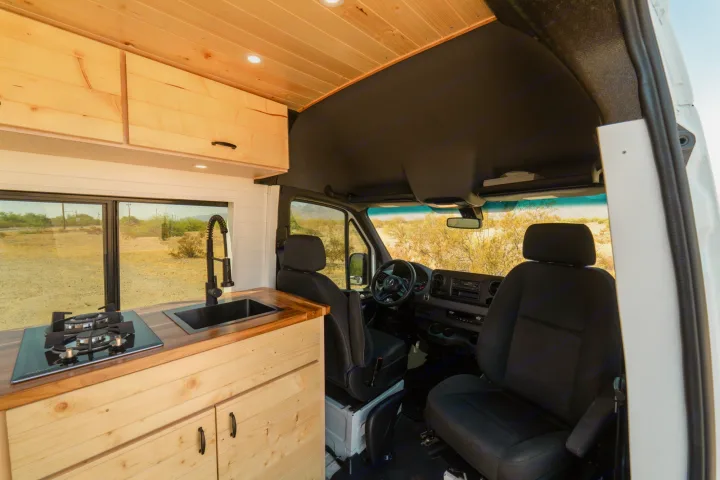 Inside this RV rental: a custom wood kitchen with sink, stove, & cabin view. Desert landscape outside.. Mercedes-Benz Sprinter 2021