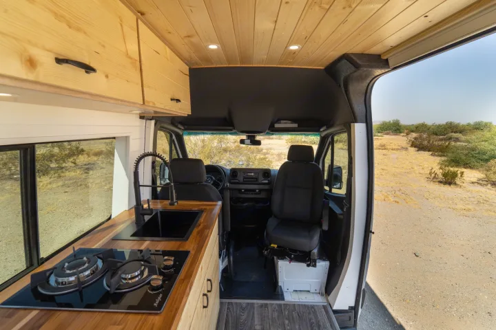 Inside an **rv rental**: light wood cabinets, ceiling, and counter. Kitchen with black sink/stove. Front seats face a desert view.. Mercedes-Benz Sprinter 2021