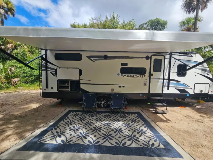 An RV rental with an extended awning, patterned outdoor rug, and two chairs at a sunny campsite.. Keystone RV Passport SL 2021
