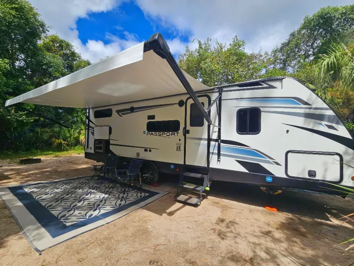This rv rental is ready for adventure! Modern travel trailer with extended awning, outdoor rug, and chairs at a scenic campsite.. Keystone RV Passport SL 2021