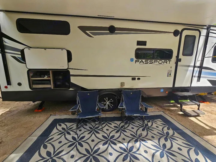 Relax in this RV rental! A travel trailer with extended awning, open storage, and two blue camp chairs on a patterned mat.. Keystone RV Passport SL 2021