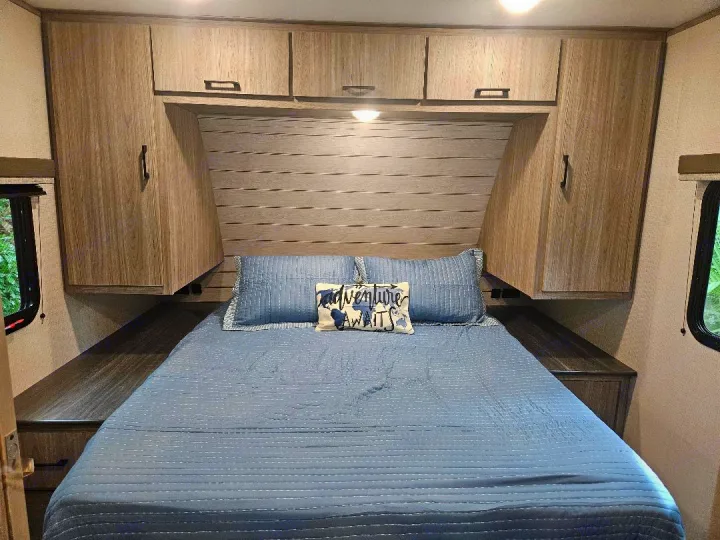 Cozy RV rental bedroom with blue bedding, overhead cabinets, and a scenic window view. Adventure awaits!. Keystone RV Passport SL 2021