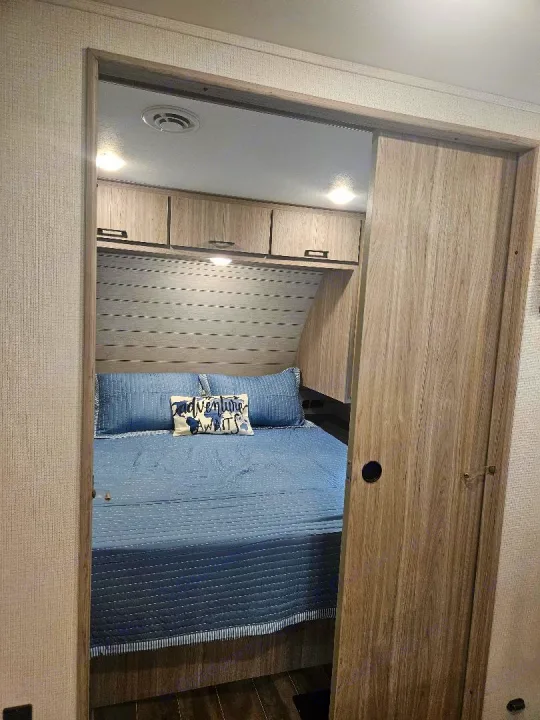 Inside an RV rental: a cozy bedroom with a blue bed, "Adventure Awaits" pillow, overhead cabinets, and wood sliding door.. Keystone RV Passport SL 2021