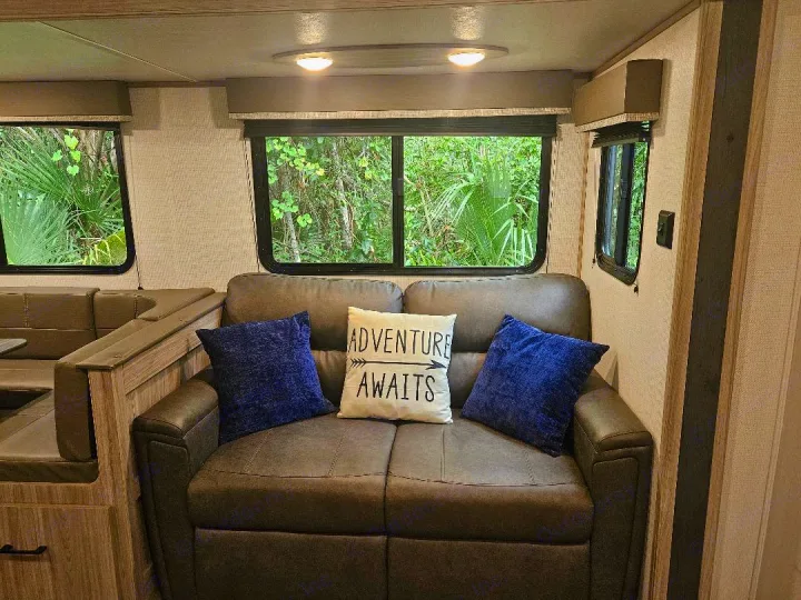 Comfortable RV rental living area with a sofa, dinette, and scenic windows revealing green outdoors. 'Adventure Awaits' pillow.. Keystone RV Passport SL 2021