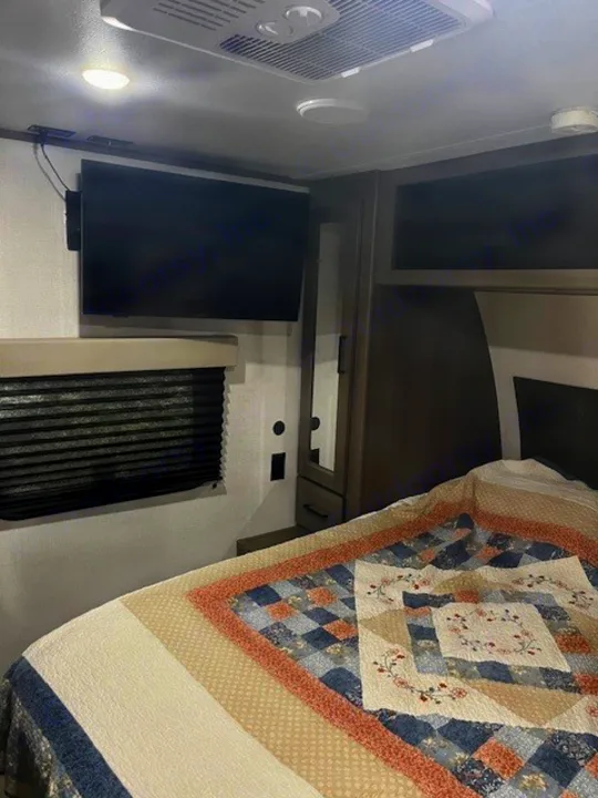 Cozy RV rental bedroom showing a bed with a patterned quilt, wall TV, and AC unit overhead.. Grand Design Transcend Xplor 2024