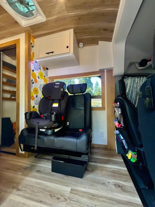 An RV rental's interior shows a child car seat on custom seating, a cabinet, and wooden floors, perfect for family adventures.. Ford Transit 2023