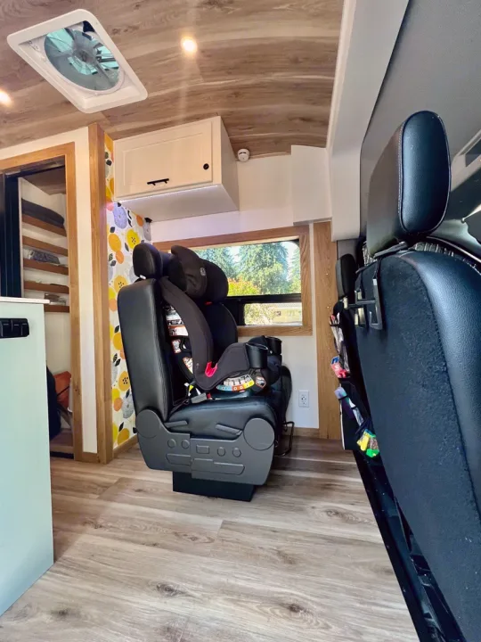 Bright rv rental interior with wood-look flooring, a child's car seat, overhead cabinets, and a ceiling vent.. Ford Transit 2023