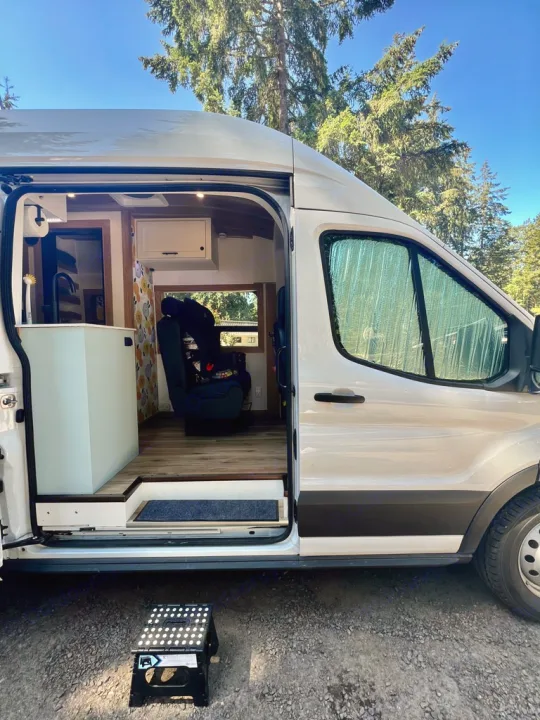Explore this white rv rental camper van! Its open side door reveals a custom interior with kitchen and seating, perfect for van life.. Ford Transit 2023