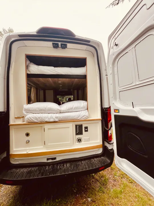 Rear view of an RV rental camper van, showing its interior with bunk beds, wooden accents, and a control panel.. Ford Transit 2023