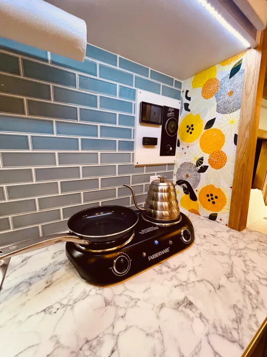 RV rental kitchen: Compact setup with Farberware cooktop, pan, kettle, blue tile backsplash, and control panel.. Ford Transit 2023