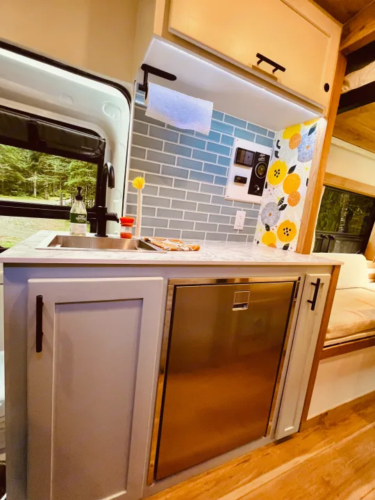 This RV rental features a modern kitchen with a sink, fridge, light-colored cabinets, and a scenic window view, perfect for van life adventures.. Ford Transit 2023