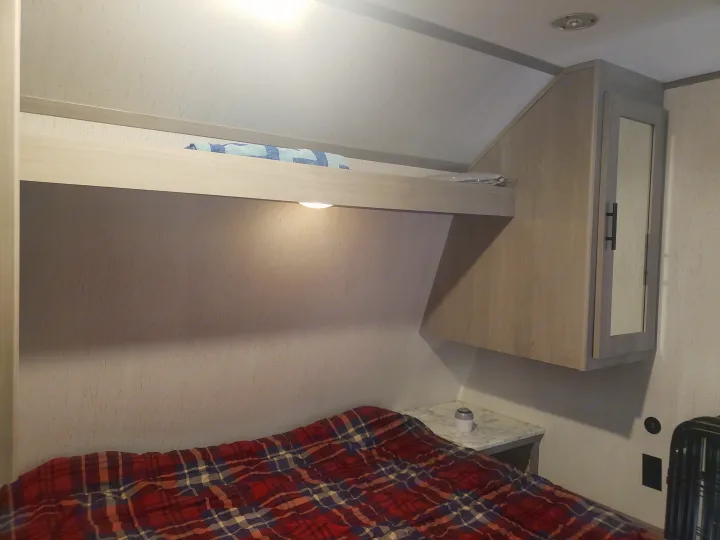 Step into this RV rental bedroom: a comfy plaid bed, overhead bunk with light, mirrored cabinet, and small nightstand.. Dutchmen Coleman Lantern LT 2022