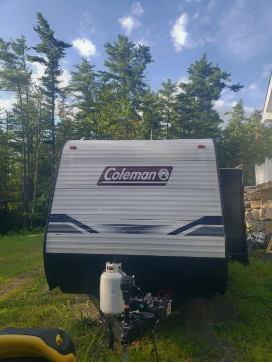 White Coleman travel trailer RV rental, with a propane tank and hitch, parked in a grassy area amidst tall trees.. Dutchmen Coleman Lantern LT 2022