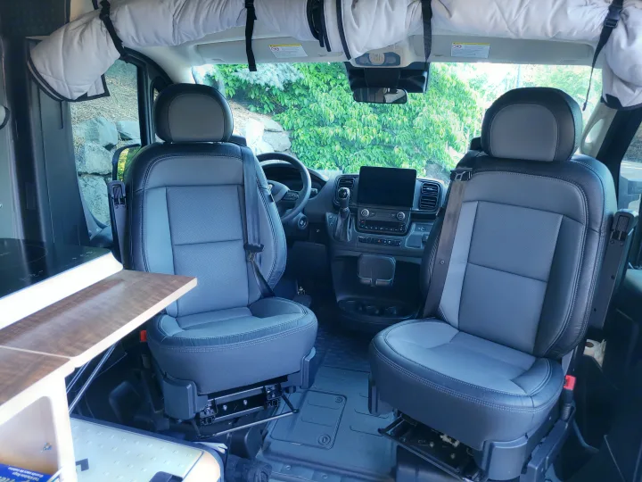 Spacious RV rental interior: front cabin with two gray seats, modern dashboard, and a side kitchenette.. Custom Ram Promaster 2023