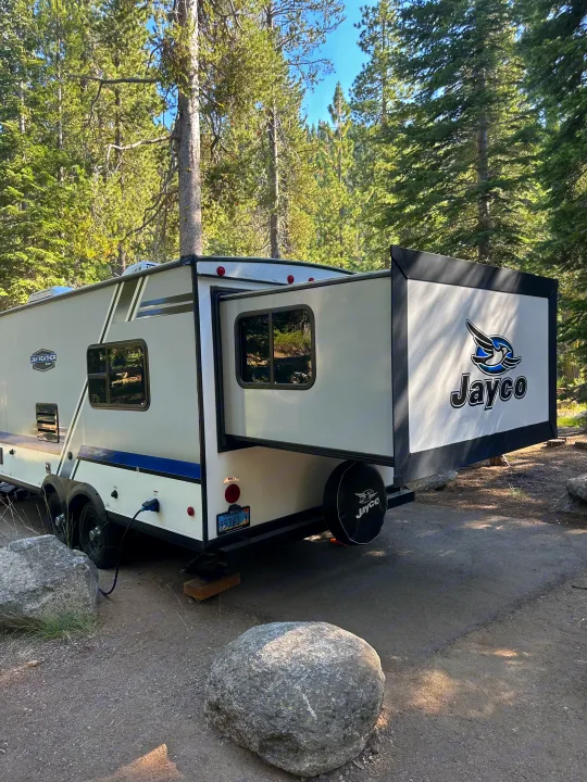 Jayco RV rental with extended pop-out parked in a serene forest campsite among tall trees.. Jayco Jay Flight 2018