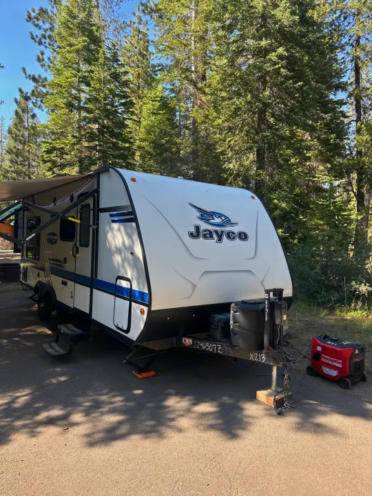 Jayco travel trailer RV rental at a sunny campsite, featuring an extended awning, generator, and tall pine trees.. Jayco Jay Flight 2018